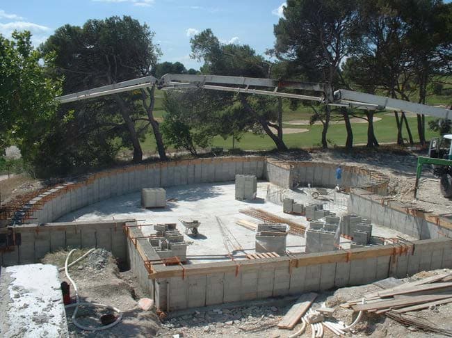 Chantier de piscine en blocs à bancher : parois montées et armées, palettes de blocs sur le radier, bras de pompe à béton.