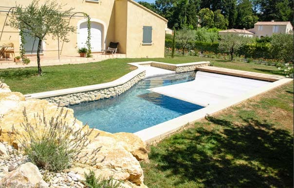 Piscine en L avec son volet de couverture, murets en pierre et maison provençale.