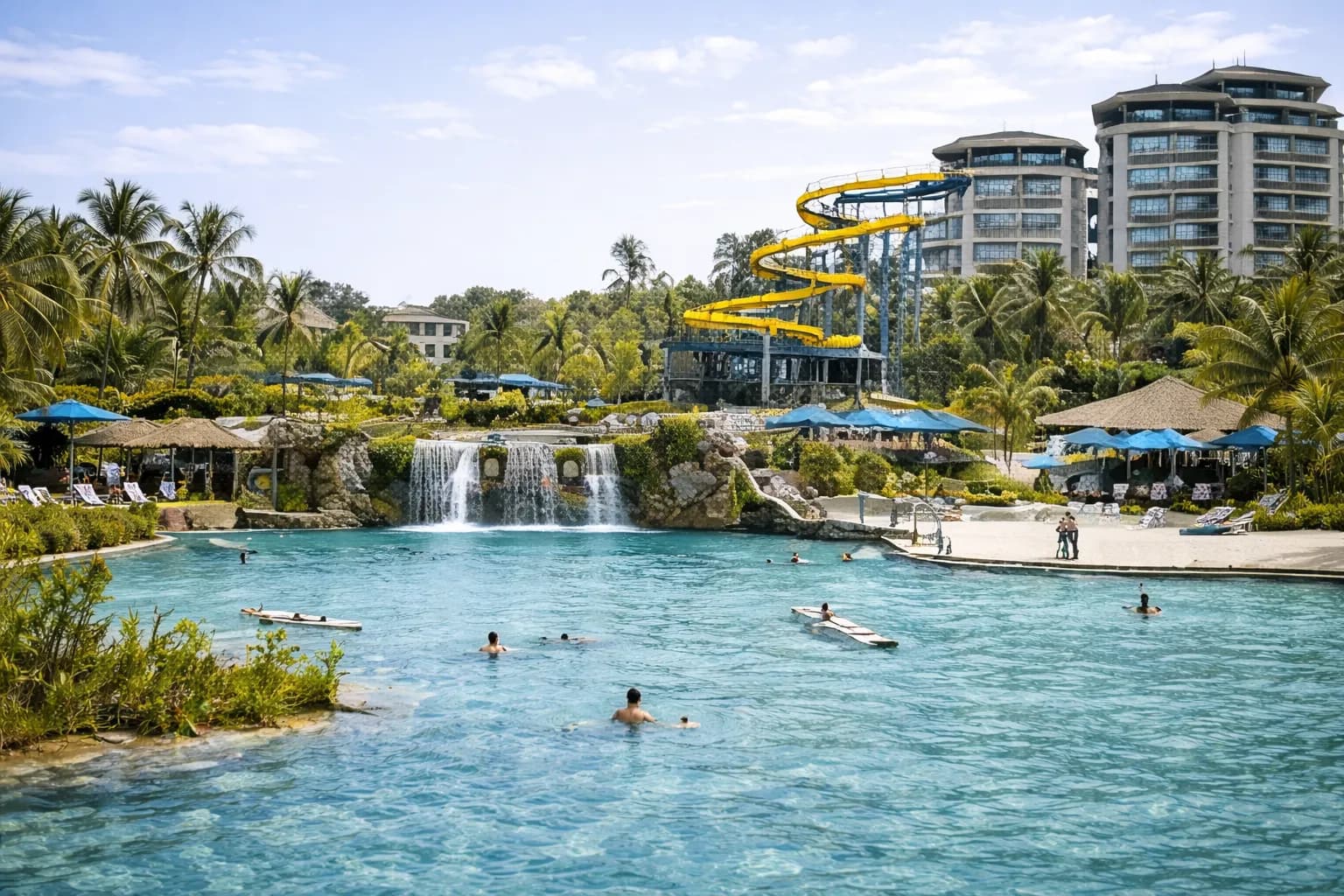 Grand bassin-lagon de resort avec cascades et toboggan, tours d'hôtel au fond.