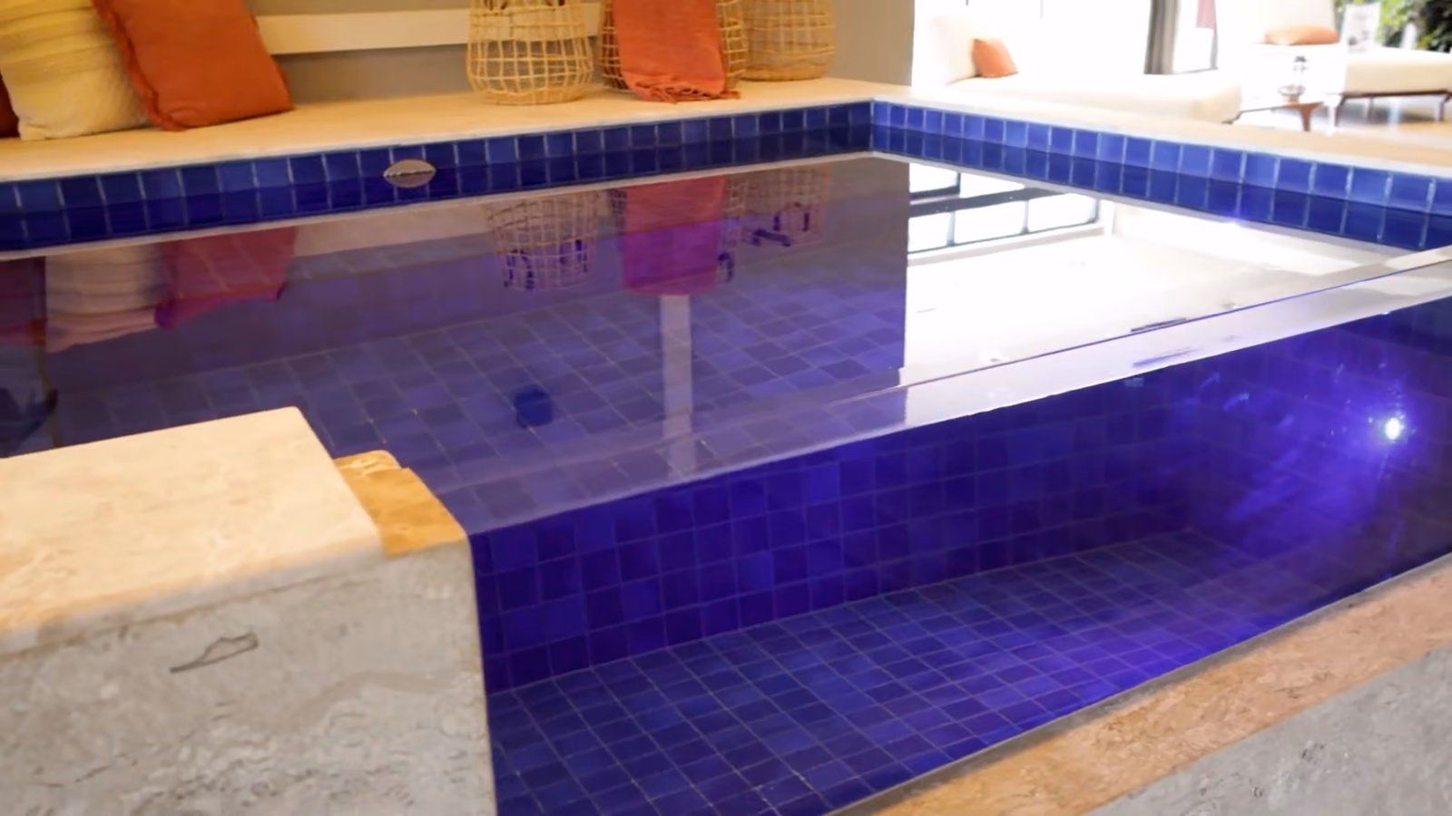 Angle d'un bassin intérieur : paroi transparente, carrelage bleu nuit, coussins orange au bord.