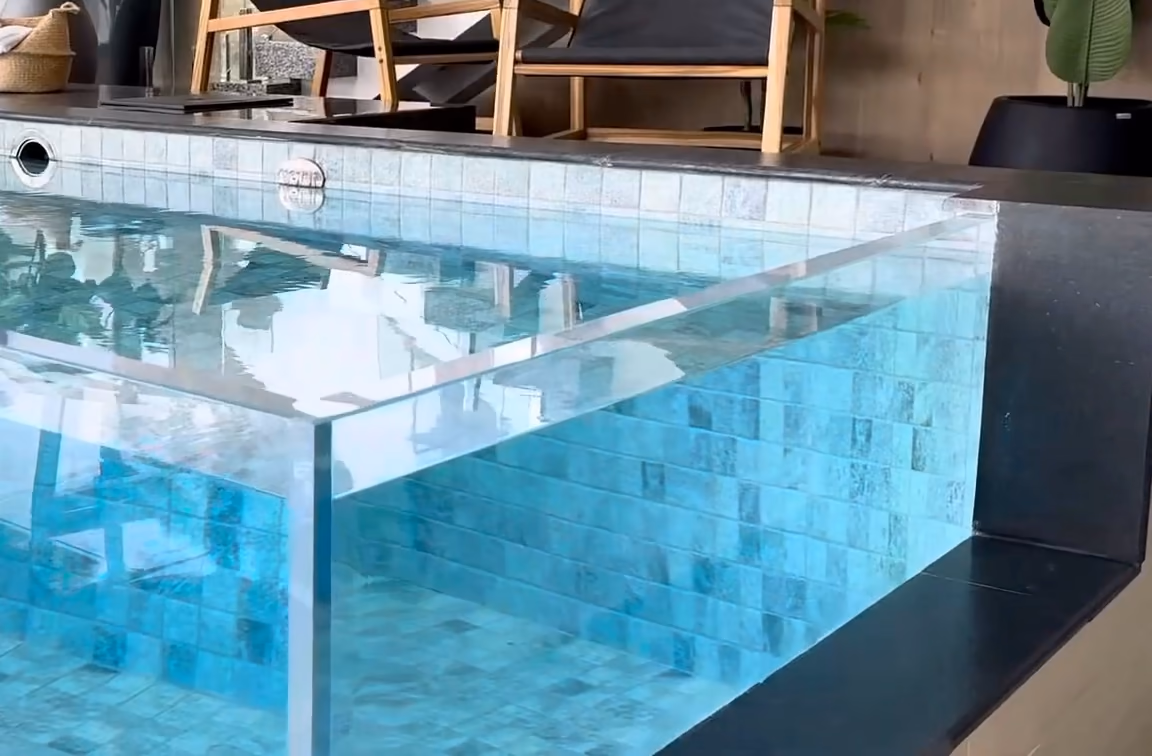 Coin d'un bassin à paroi transparente sur une terrasse : eau claire, carrelage turquoise, transats en bois.