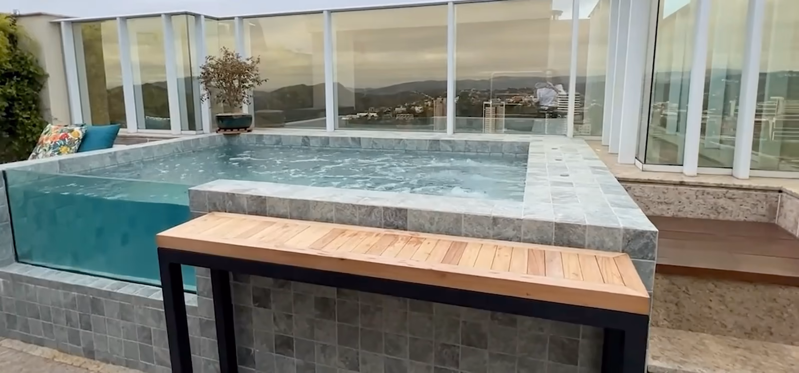 Toit-terrasse fermé de garde-corps vitrés : un bassin à paroi transparente, un banc en bois, la ville au loin.
