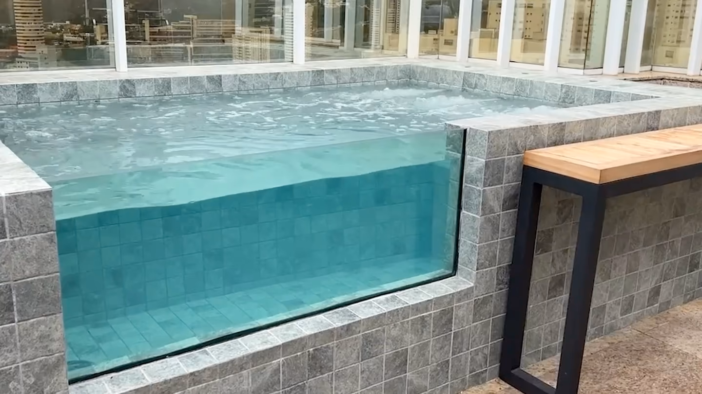 Paroi transparente d'un bassin sur un toit-terrasse, eau turquoise, banc en bois sur le côté.