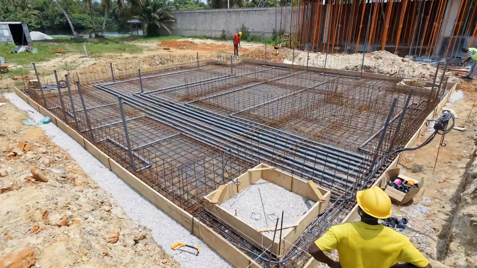 Sur le chantier Villa Park, un ouvrier casqué, de dos, devant le radier ferraillé ; un autre ouvrier au fond, près de la structure sous étais.