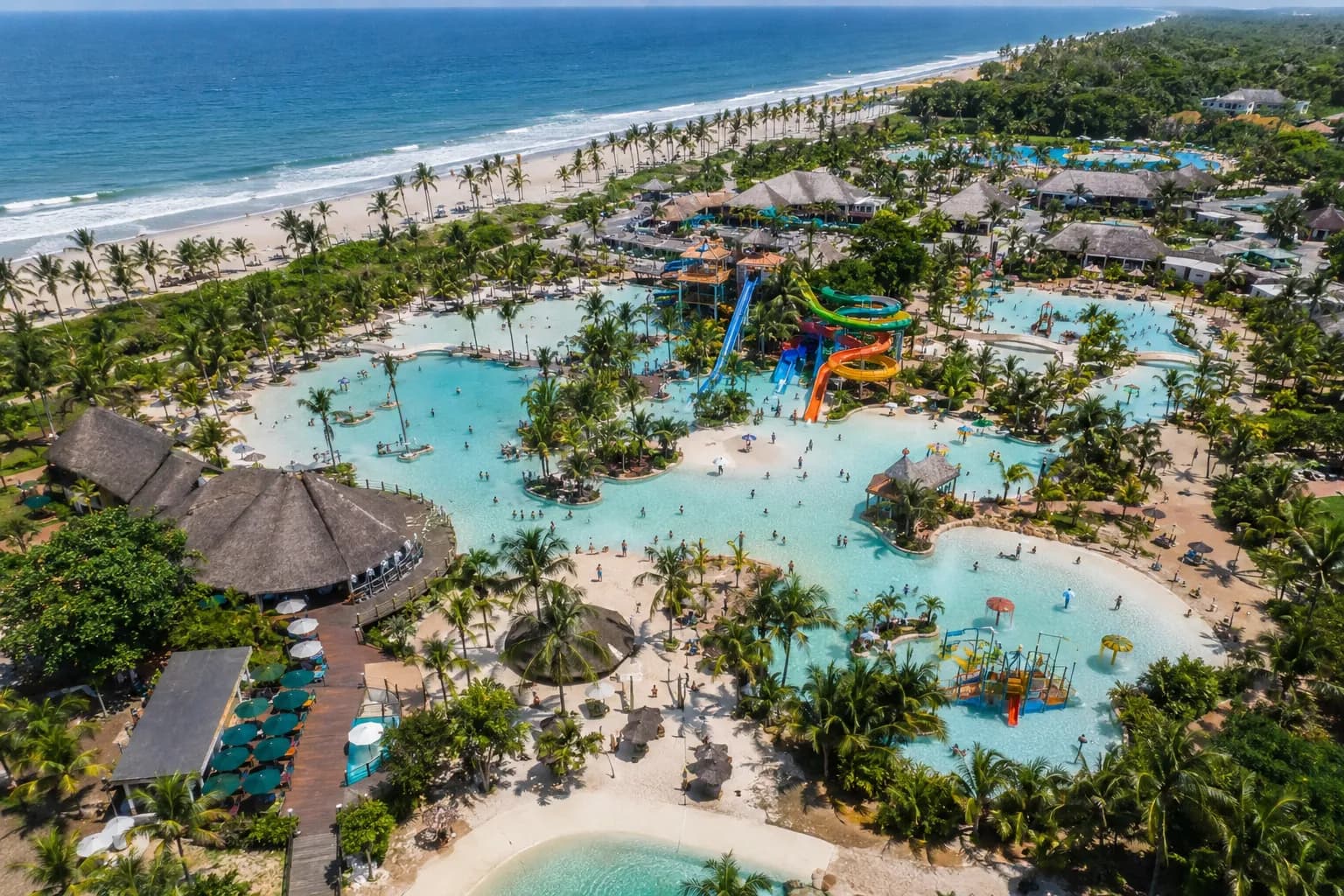 Parc aquatique tropical en bord d'océan vu du ciel : lagons, toboggans, paillotes, plage.