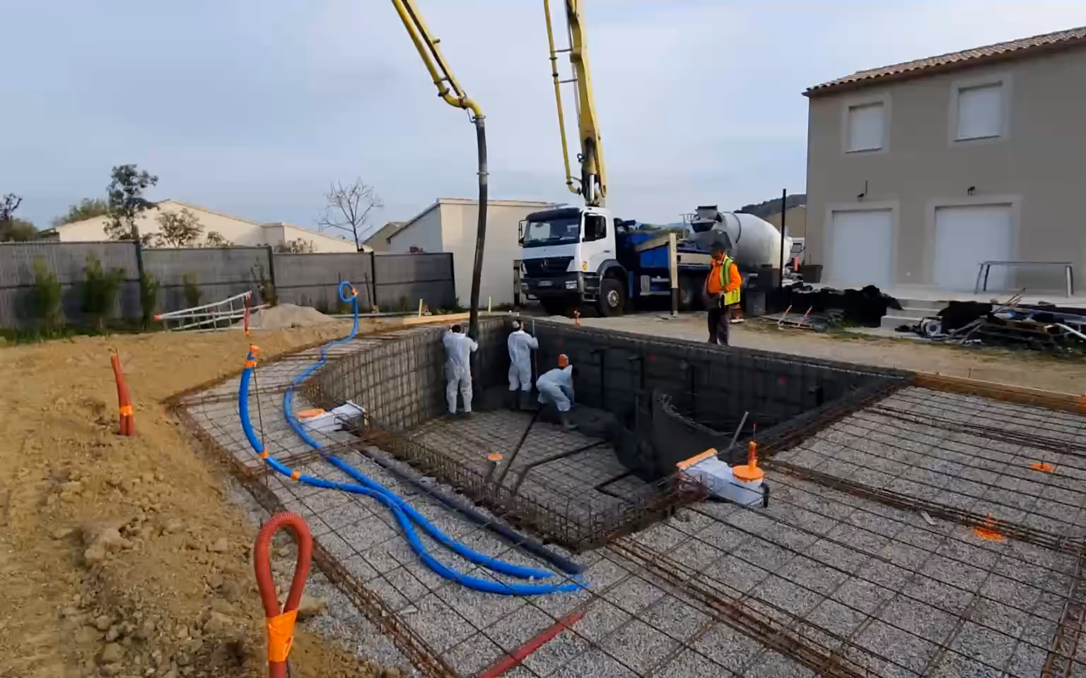 Chantier d'une piscine en béton armé en France : pompe à béton au-dessus du bassin ferraillé, béton frais contre le treillis, radier tiré, puis la coque coulée.