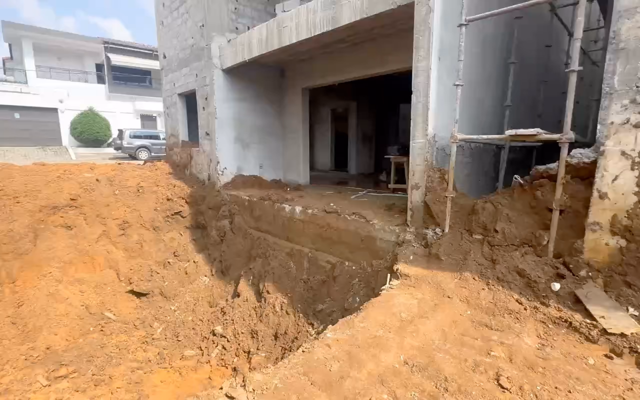 Terrassement sur notre chantier d'Abidjan : une fouille en terre orange le long d'un rez-de-chaussée en gros œuvre, puis le talus de terre rouge.