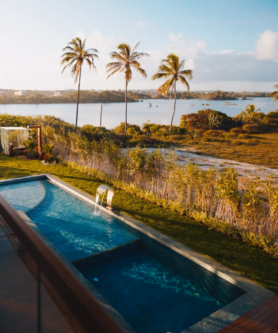 Depuis l'étage d'une villa, la piscine, le jardin et une lagune bordée de cocotiers au soleil couchant.