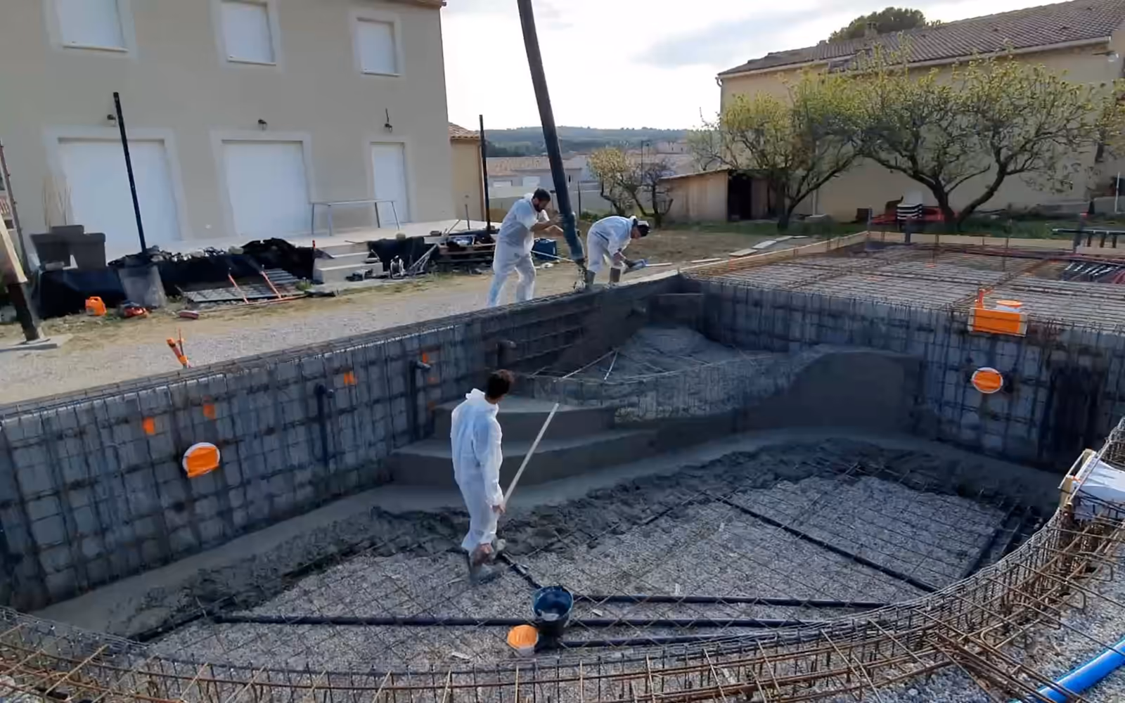 Coulage d'une piscine monobloc de forme libre en France : un compagnon tire le béton du fond au pied des marches, l'équipe coule les marches romaines, puis le bassin coulé et sa plage.