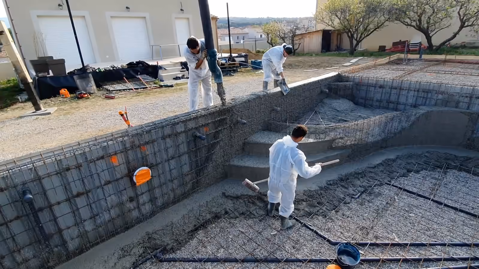 Chantier d'une piscine monobloc en France : des compagnons tirent le béton frais au râteau au pied des marches du bassin.
