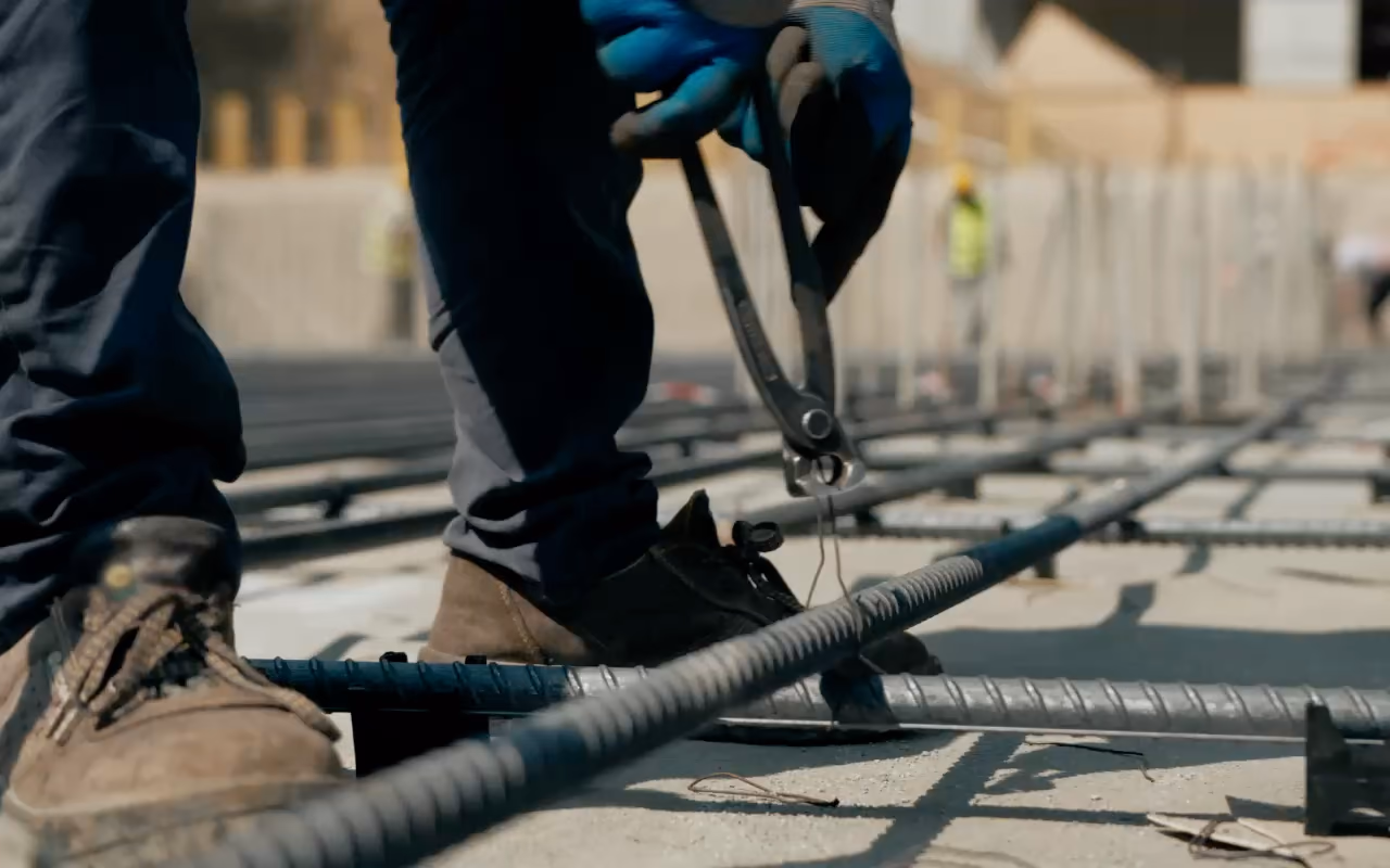 Au ras d'une nappe de ferraillage, des mains gantées ligaturent les barres, puis le béton frais est coulé et tiré au sol.