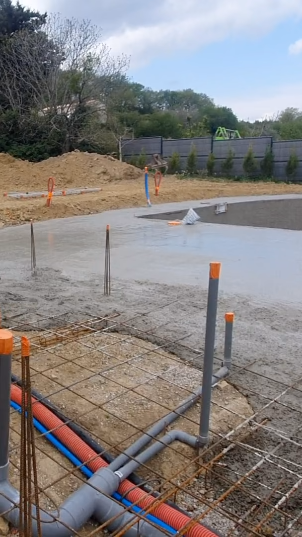 Avant le coulage d'une dalle, des gaines rouges et bleues et des tuyaux gris posés en attente dans le ferraillage, le béton frais au fond.