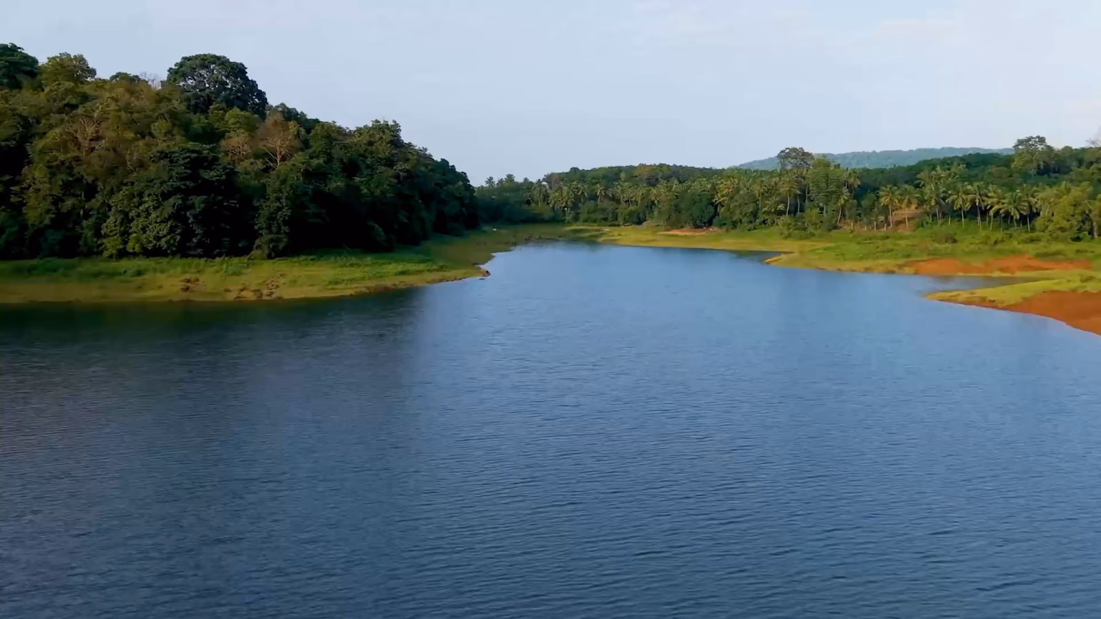 Vue de drone qui avance lentement au-dessus d'un plan d'eau calme, bordé de forêt et de rives en terre rouge.