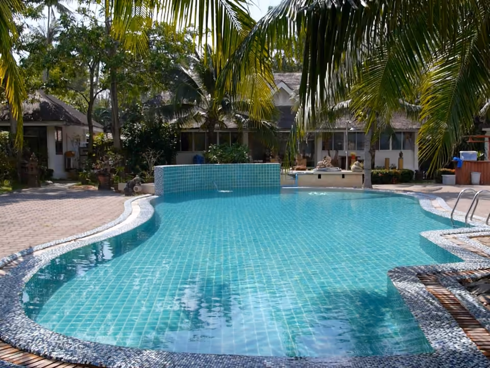 Piscine d'hôtel en mosaïque turquoise, de forme libre, sous les cocotiers, bungalows au fond.