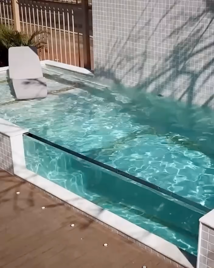 Bassin adossé à un mur carrelé, paroi transparente côté terrasse, ombres d'arbre sur l'eau.