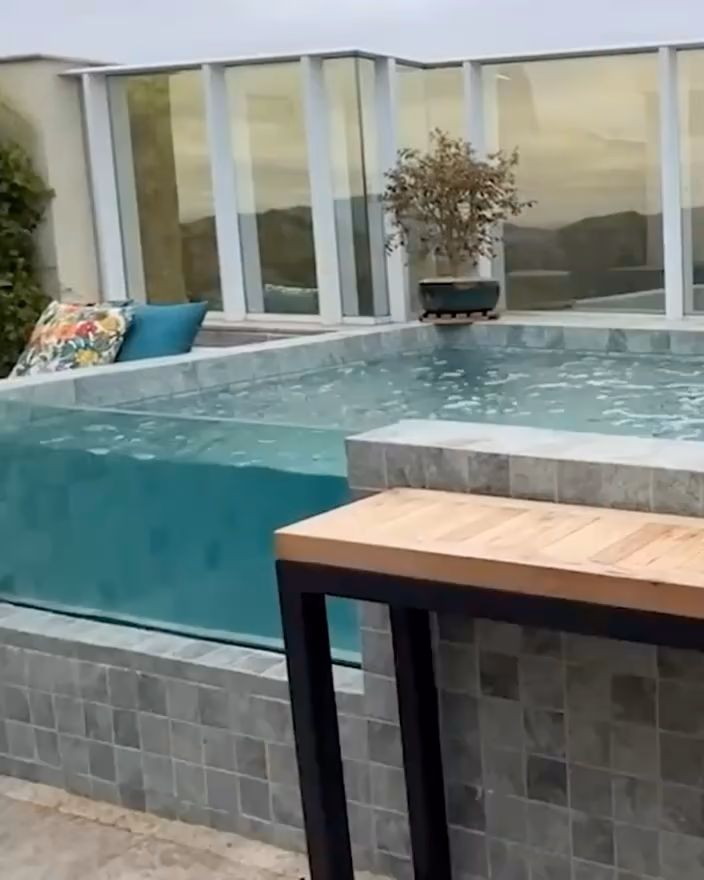 Bassin à paroi transparente sur un toit-terrasse, un banc en bois devant, garde-corps vitré et collines au loin.