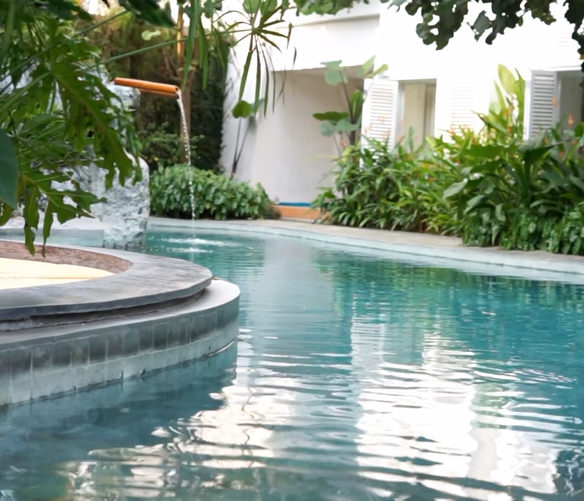 Lagon aux bords courbes en pierre, eau turquoise et petite cascade dans un jardin tropical.