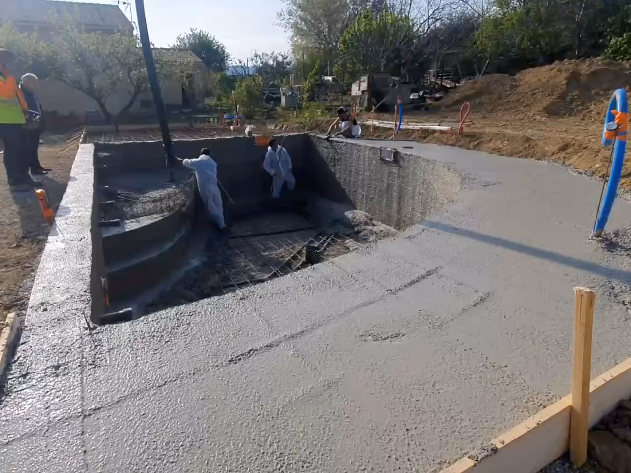 Piscine en béton armé de forme libre : la plage tout juste coulée épouse la courbe du bassin, puis les marches et le voile courbe ferraillés.