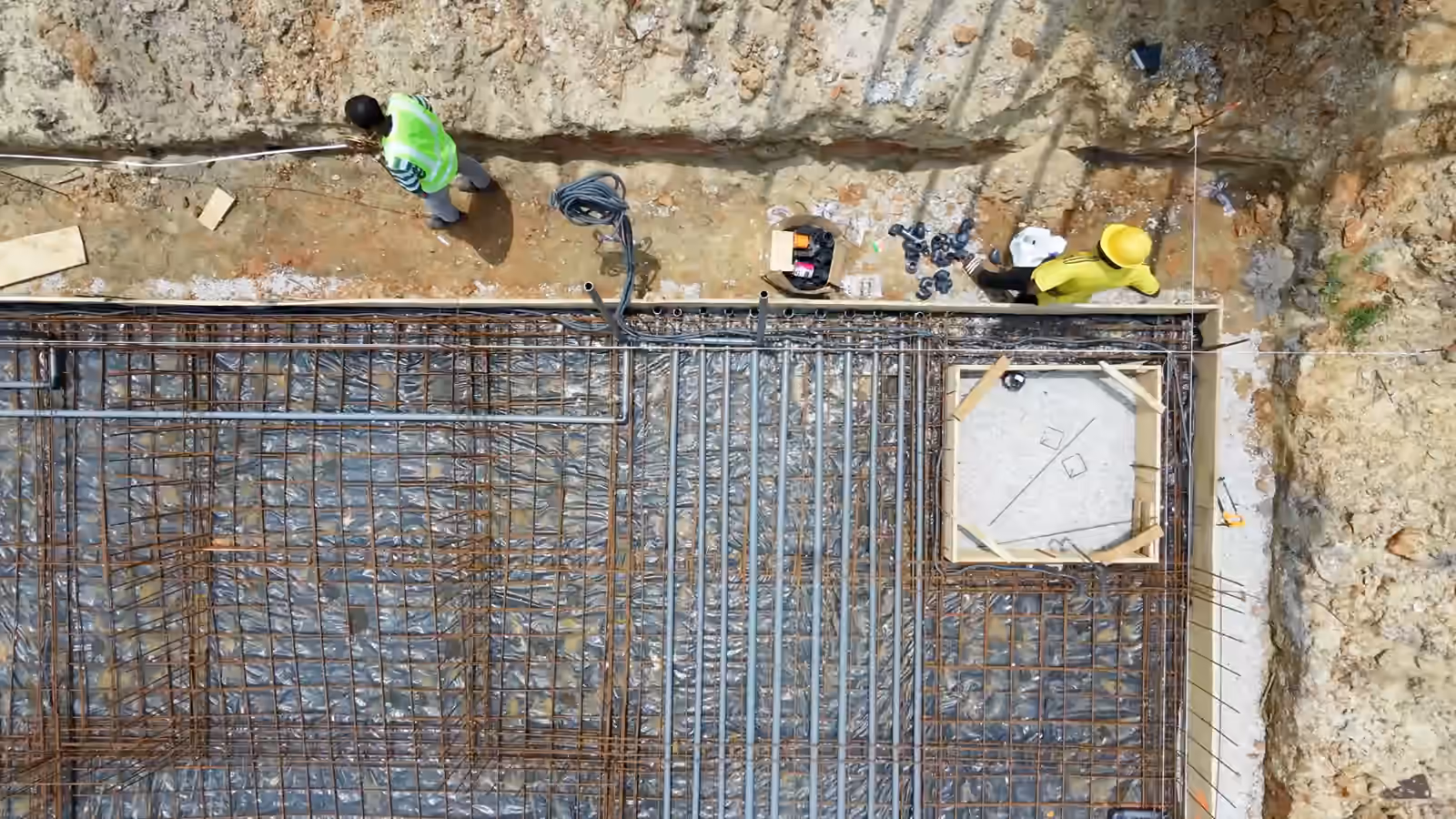 Vue verticale de drone sur le chantier Villa Park : des ouvriers le long du coffrage, au bord du radier ferraillé.