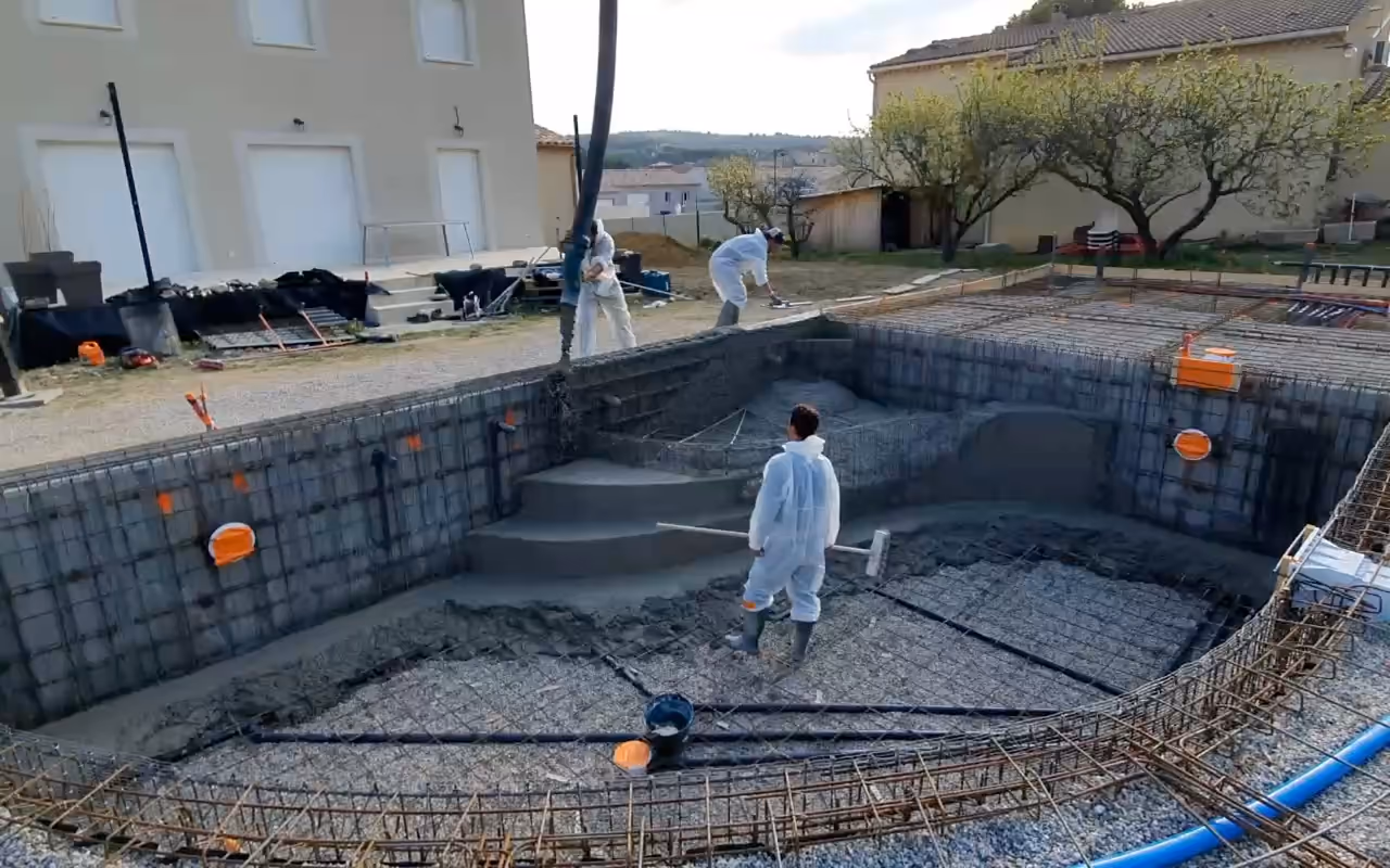 Coulage du radier d'une piscine monobloc en béton armé : compagnons en combinaison blanche, armatures et murs déjà coulés.