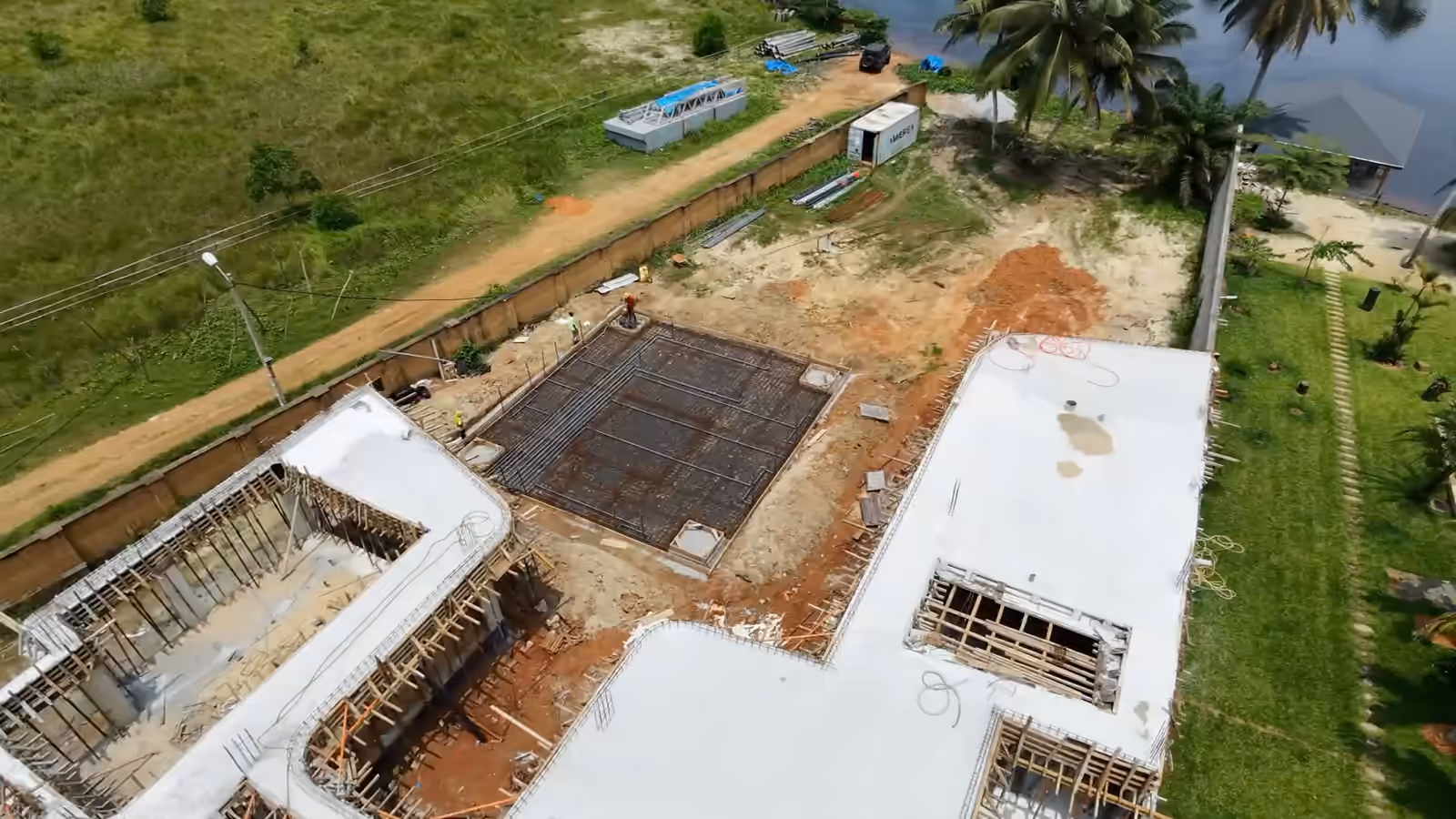 Vue de drone qui tourne autour du chantier Villa Park : la structure en béton sous étais, le radier ferraillé, la lagune au fond.