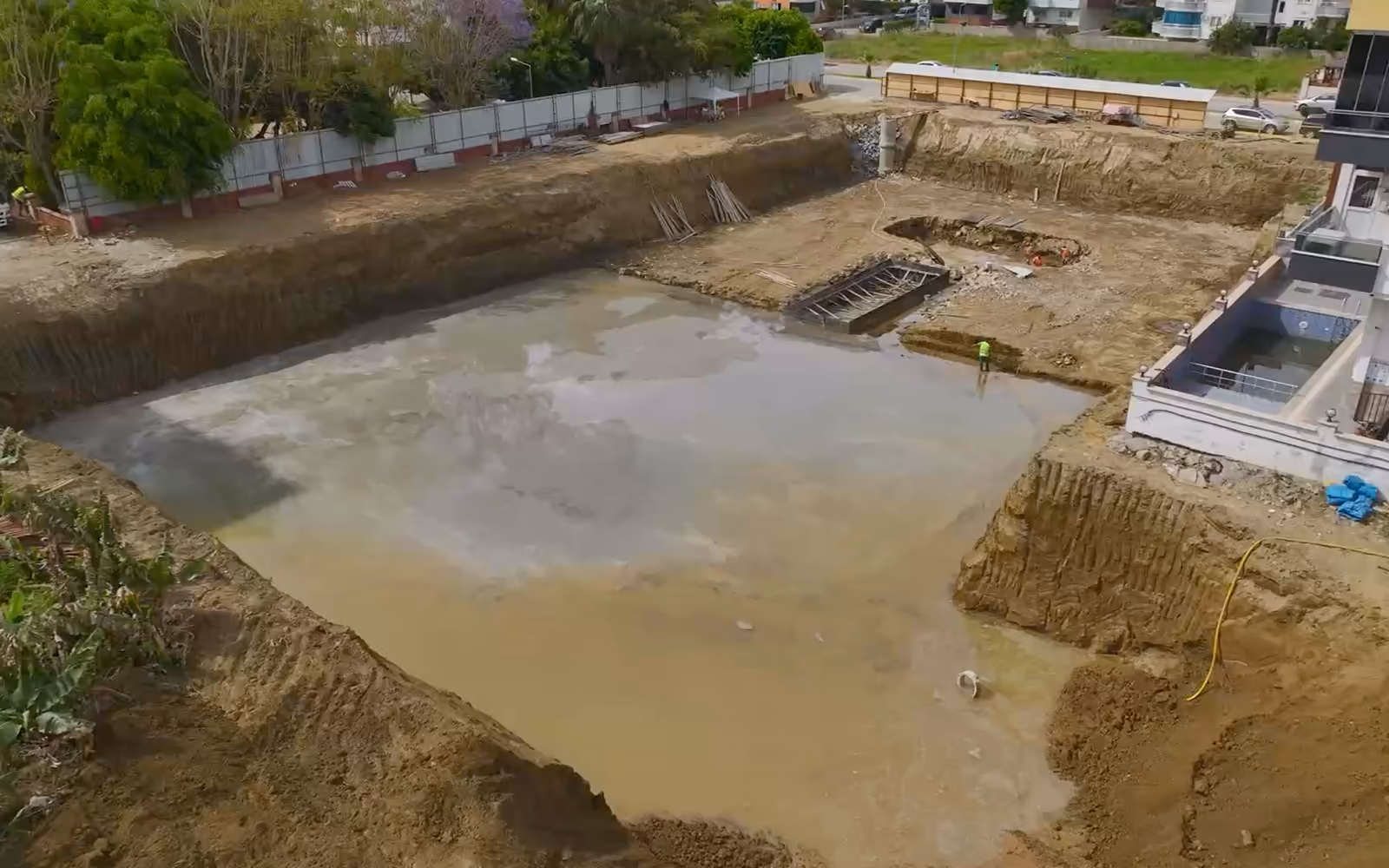 Grande fouille de chantier en ville vue du ciel : fond nivelé, talus de terre, immeubles et maisons autour.