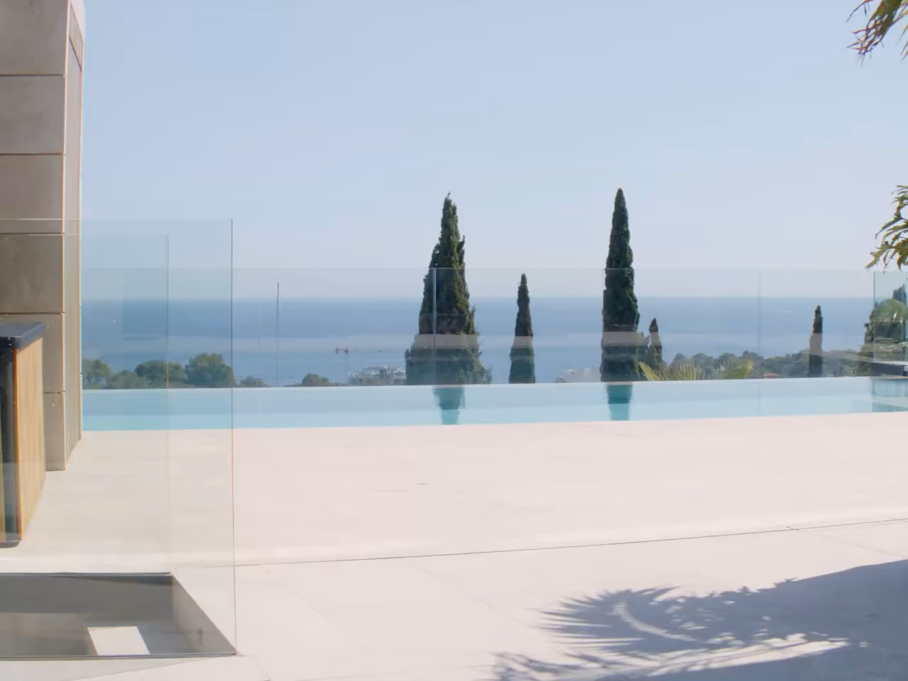 Patio d'une villa contemporaine : dallage clair, piscine à débordement, cyprès et mer au loin.