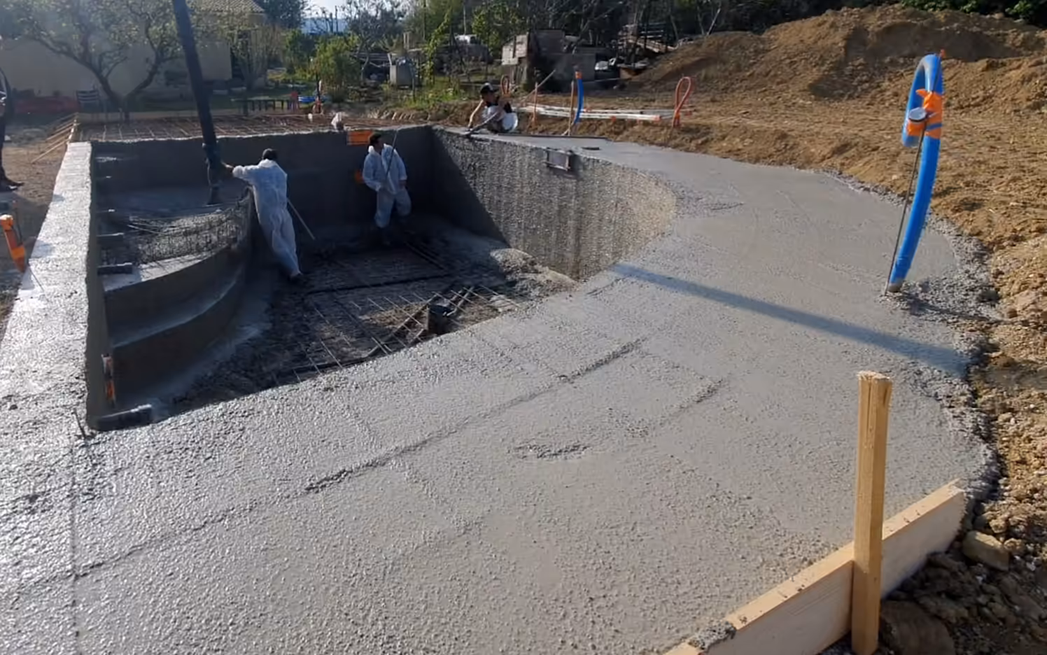 Plage en béton frais qui suit la courbe d'un bassin de forme libre, coffrage en bois et piquets, compagnons au fond.