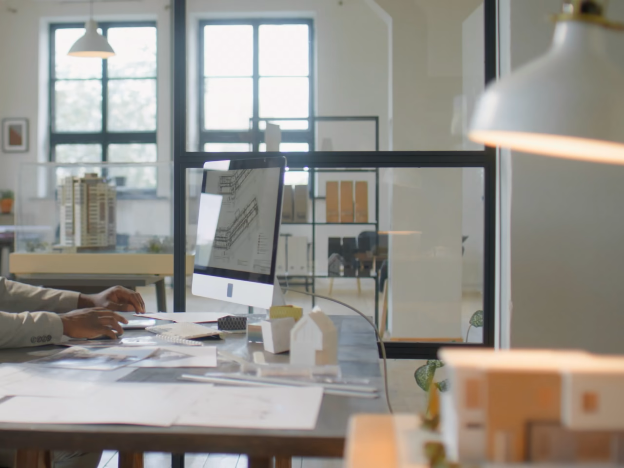 Bureau d'études : un plan affiché à l'écran, maquettes de maisons sur la table de travail.